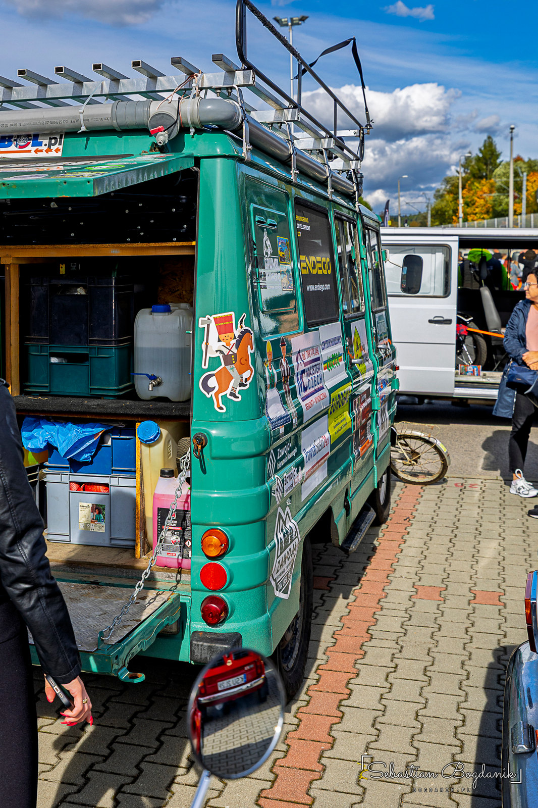 A mint-green camper with a roof rack, shelves, crates and a water jug