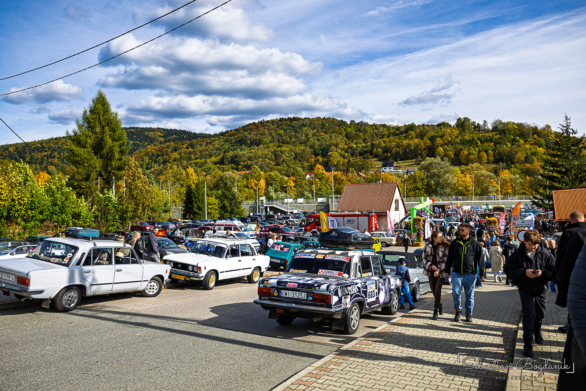 Rows of classic cars and people strolling across the square, wooded hills in autumn colours behind.