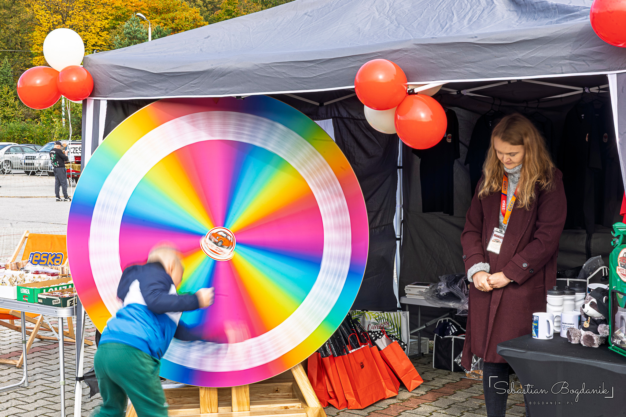 A child spins a rainbow wheel of fortune under the stand's tent, with red and white balloons nearby.