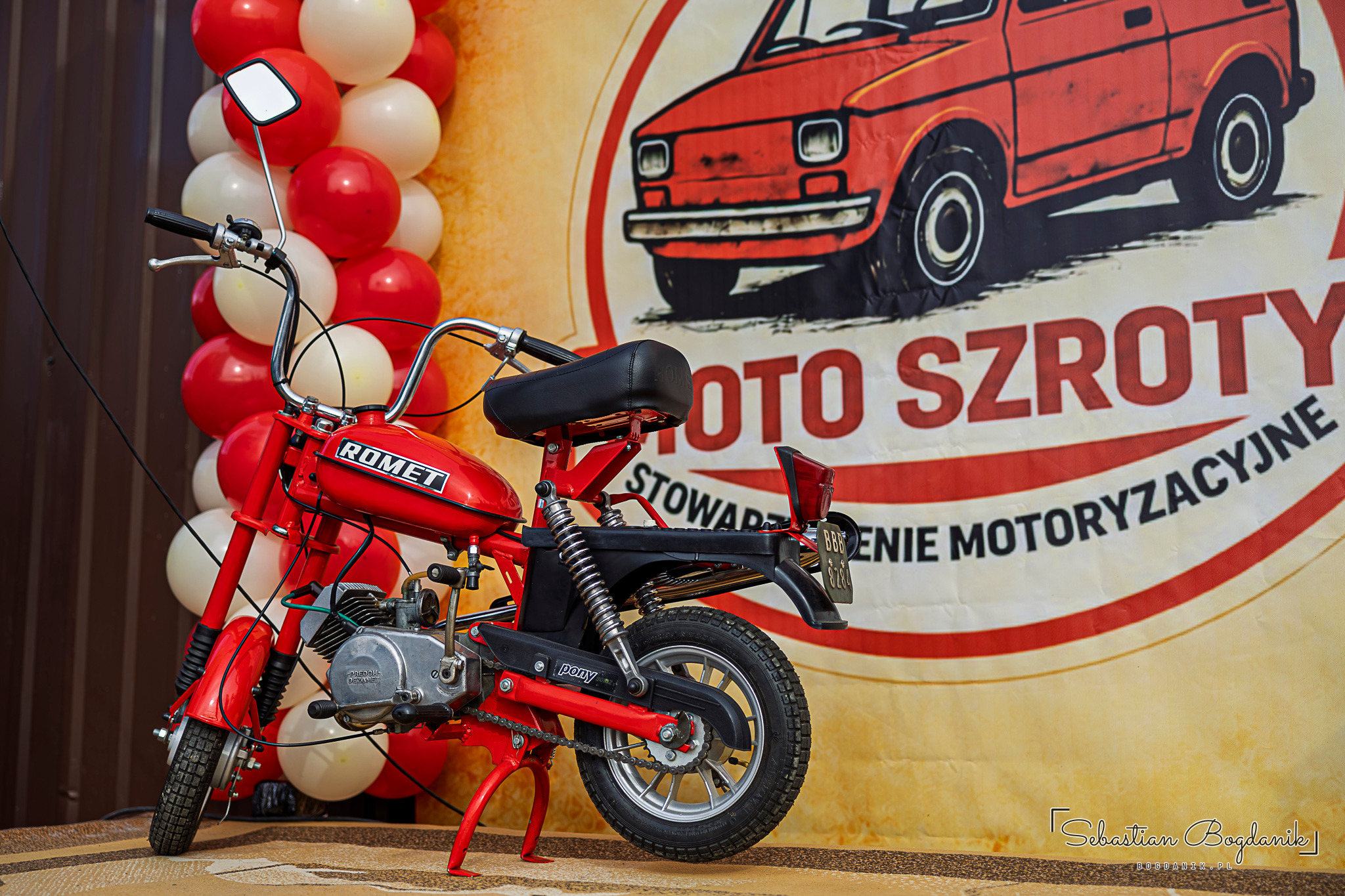 A red Romet moped in front of a banner with a drawing of a Fiat 126p and the words Moto Szroty.