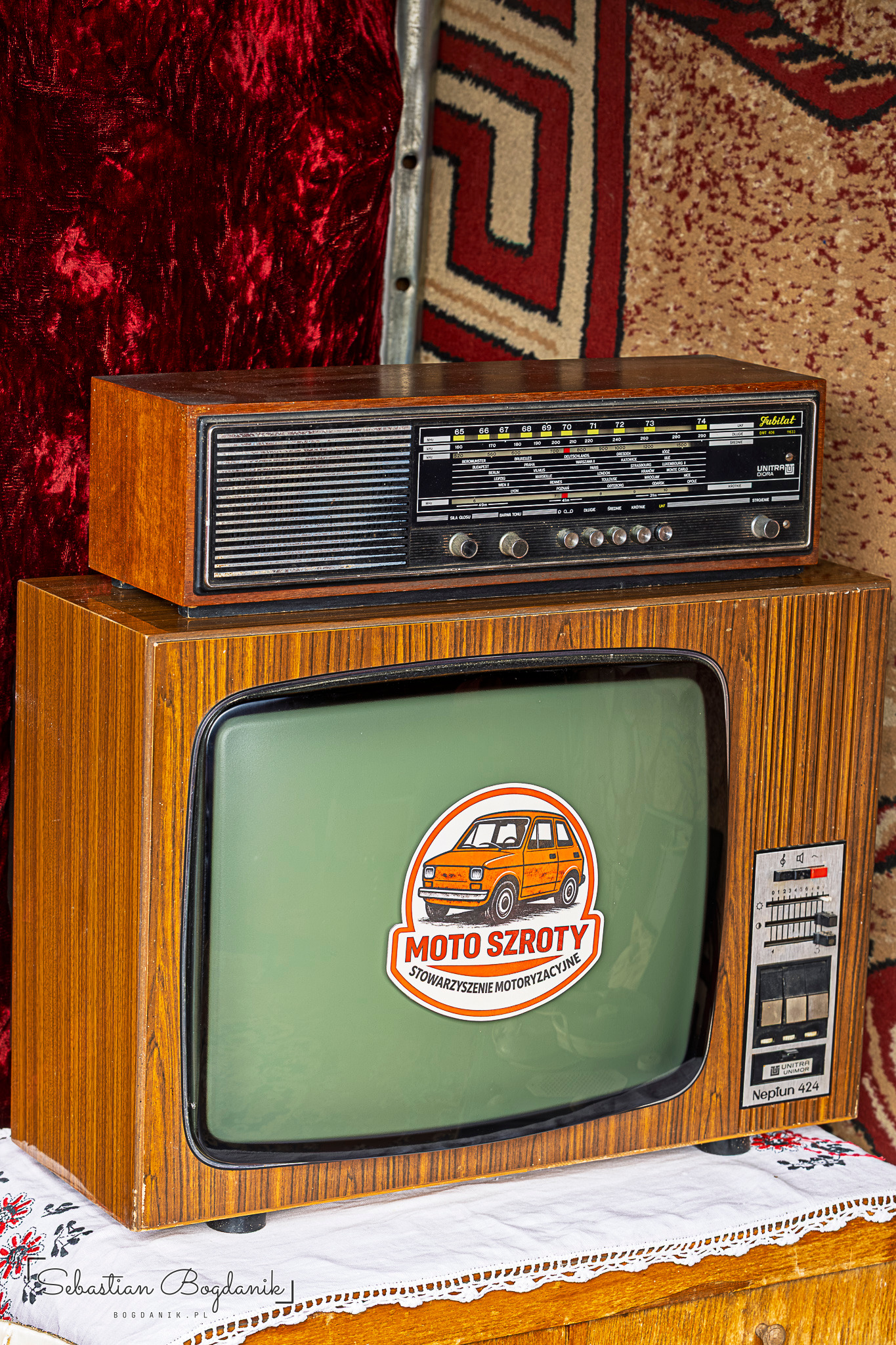 A wooden TV set with a Moto Szroty sticker on the screen, a radio on top and a patterned rug behind.
