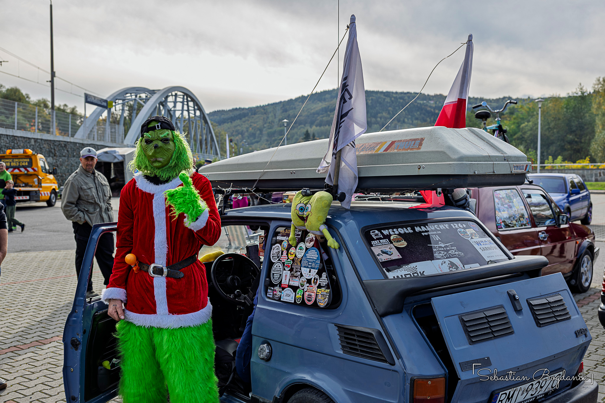 A person in a Grinch costume beside a blue Fiat 126p with a roof rack and stickers on the windows.