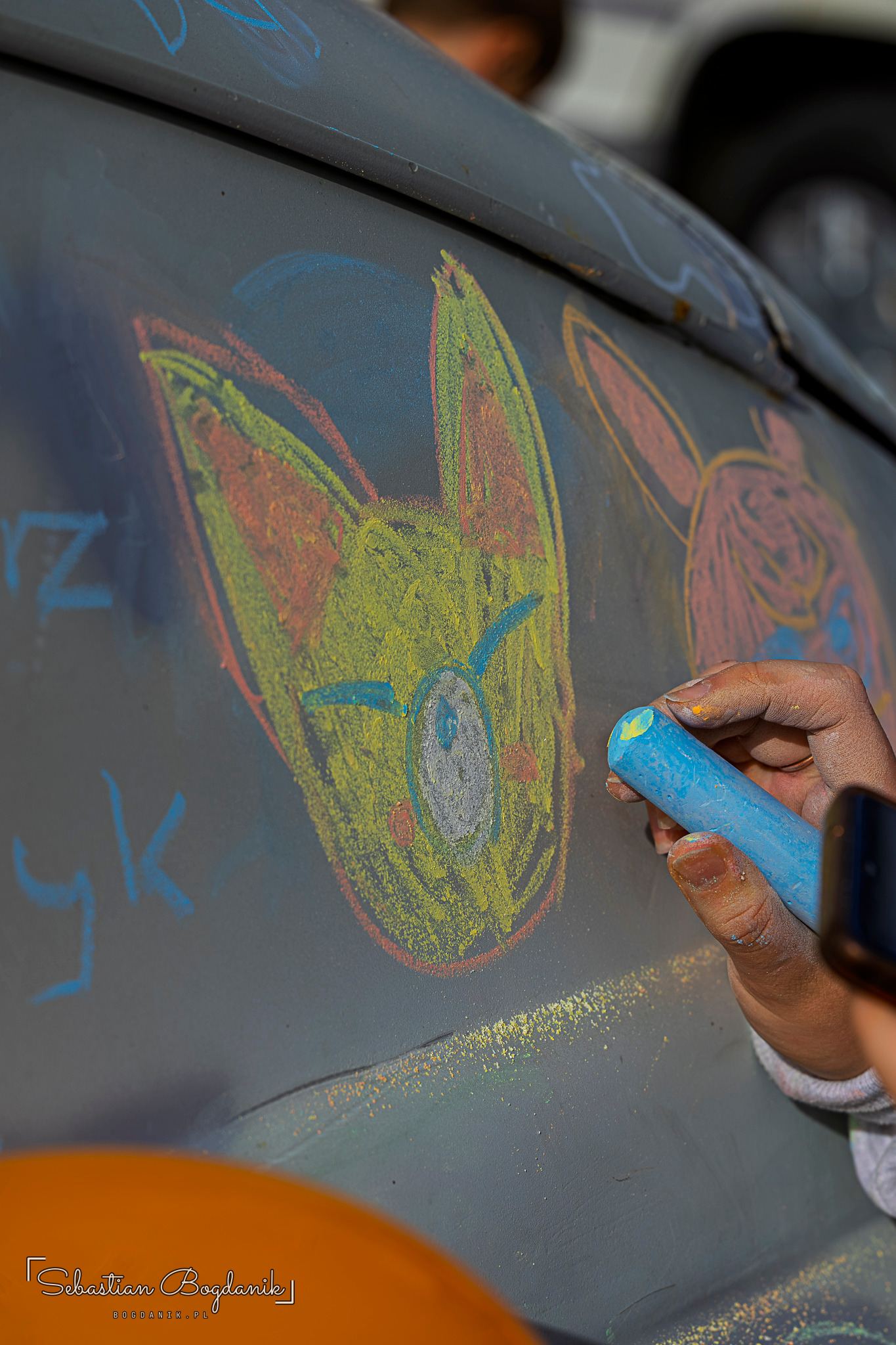 A hand holding blue chalk draws colourful figures on the grey body of a car.