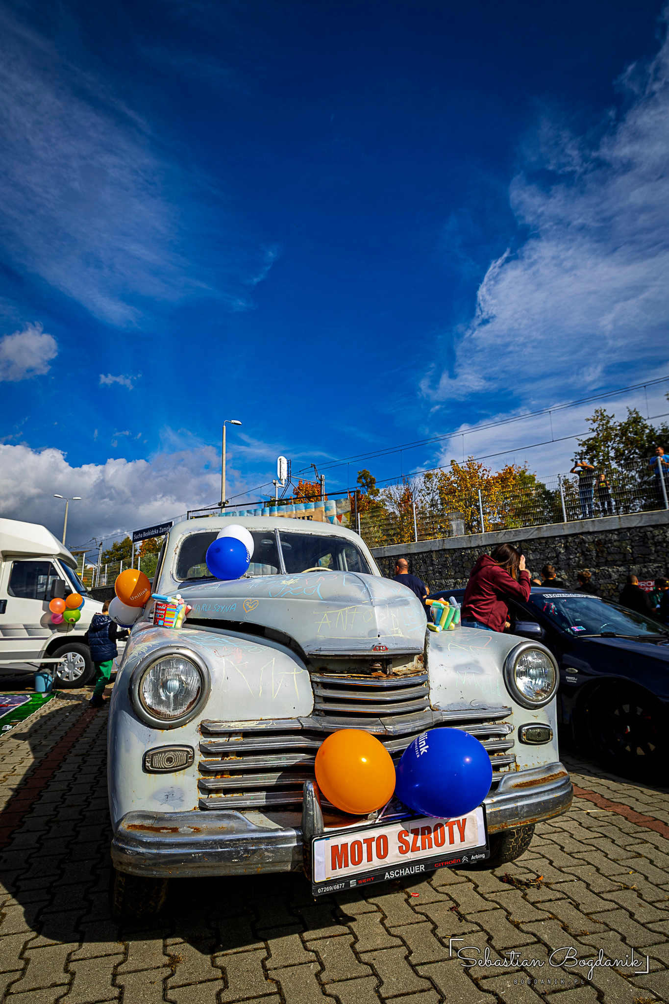 A white Pobeda with a chrome grille and colourful balloons on the bumper, under a blue sky.