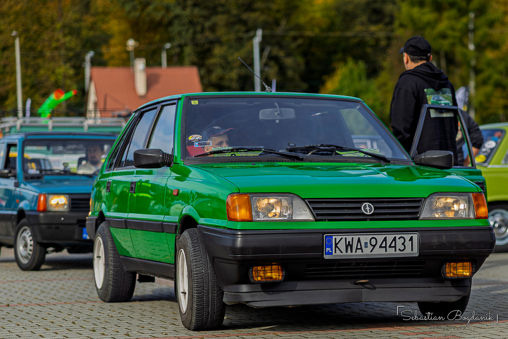 A green Polonez Caro driving onto the square, other classic cars behind it.