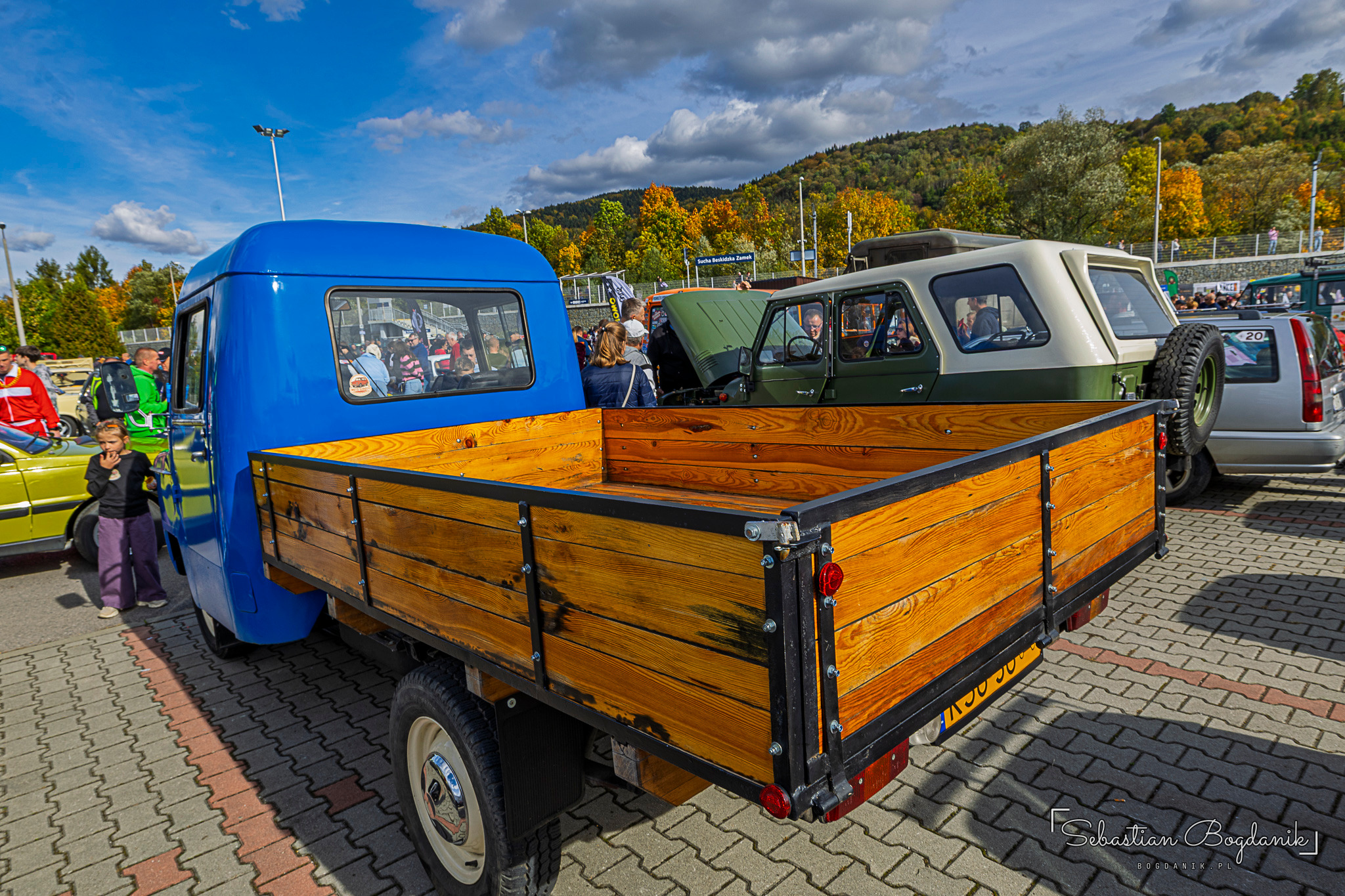 The blue cab of a Nysa with an empty wooden load bed, other cars and autumn hills behind.