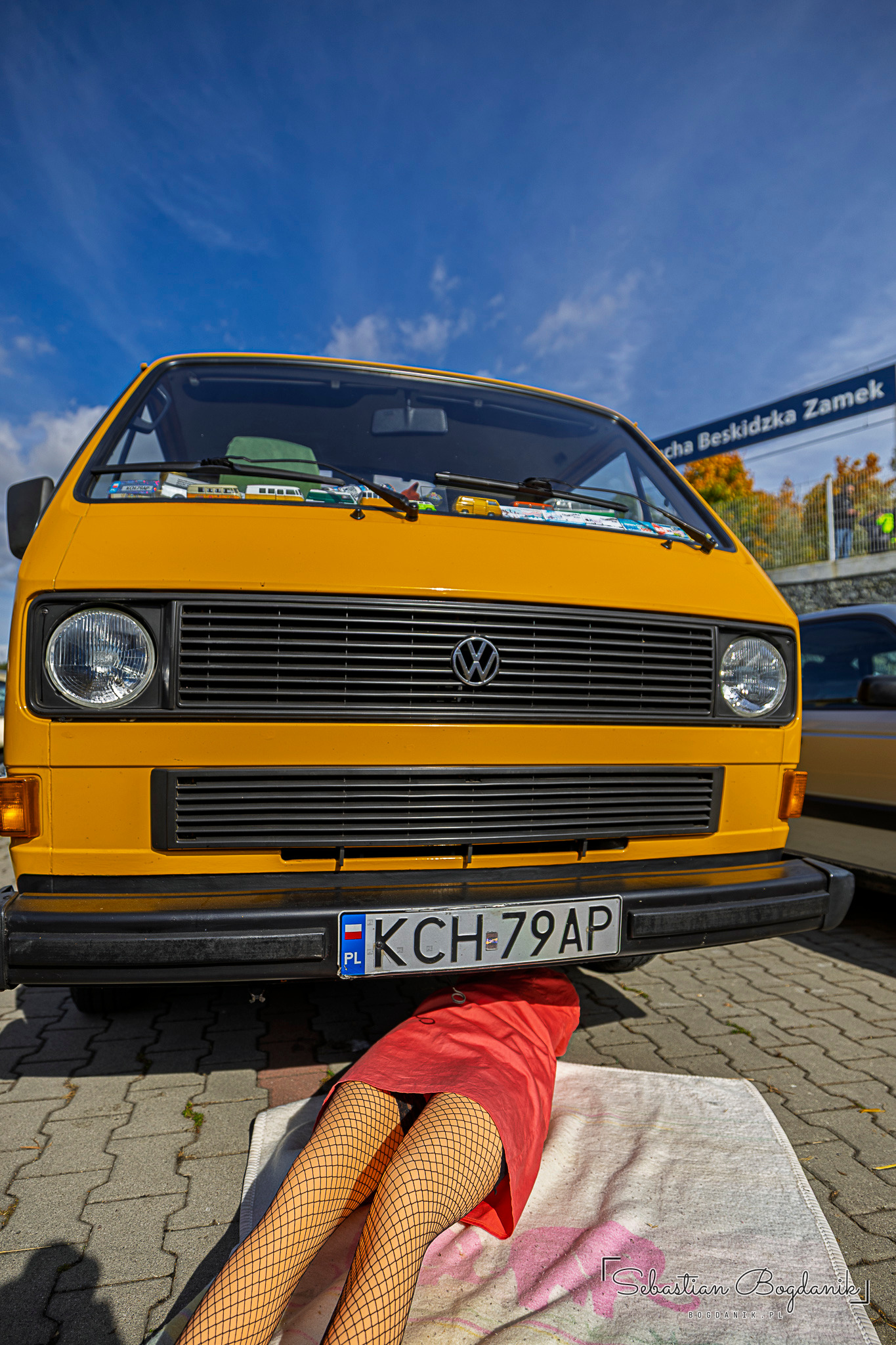 A yellow Volkswagen T3 from the front, with a mannequin's legs in fishnet tights and a red dress sticking out from under the bumper.