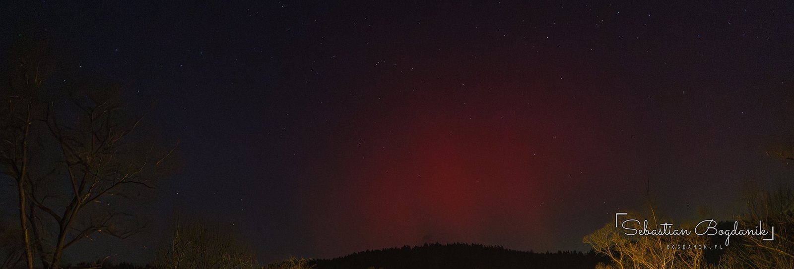 22:28. Czerwona kolumna światła nad lasem.